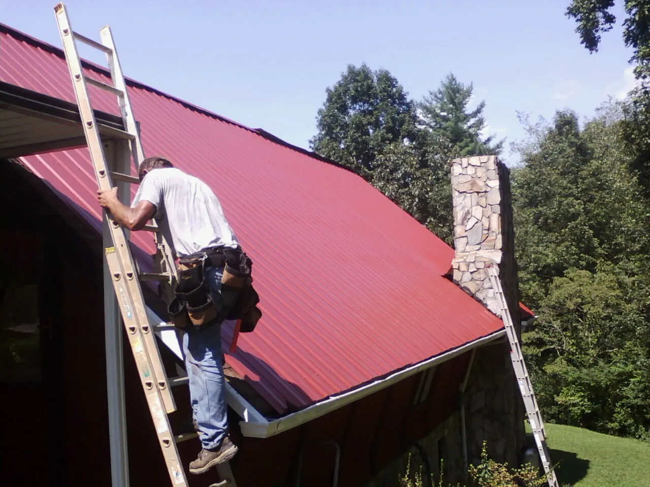 Roofer on a ladder beside a red metal roof and stone chimney — Soffit & Fascia Repair by Roofing Brothers in Peru